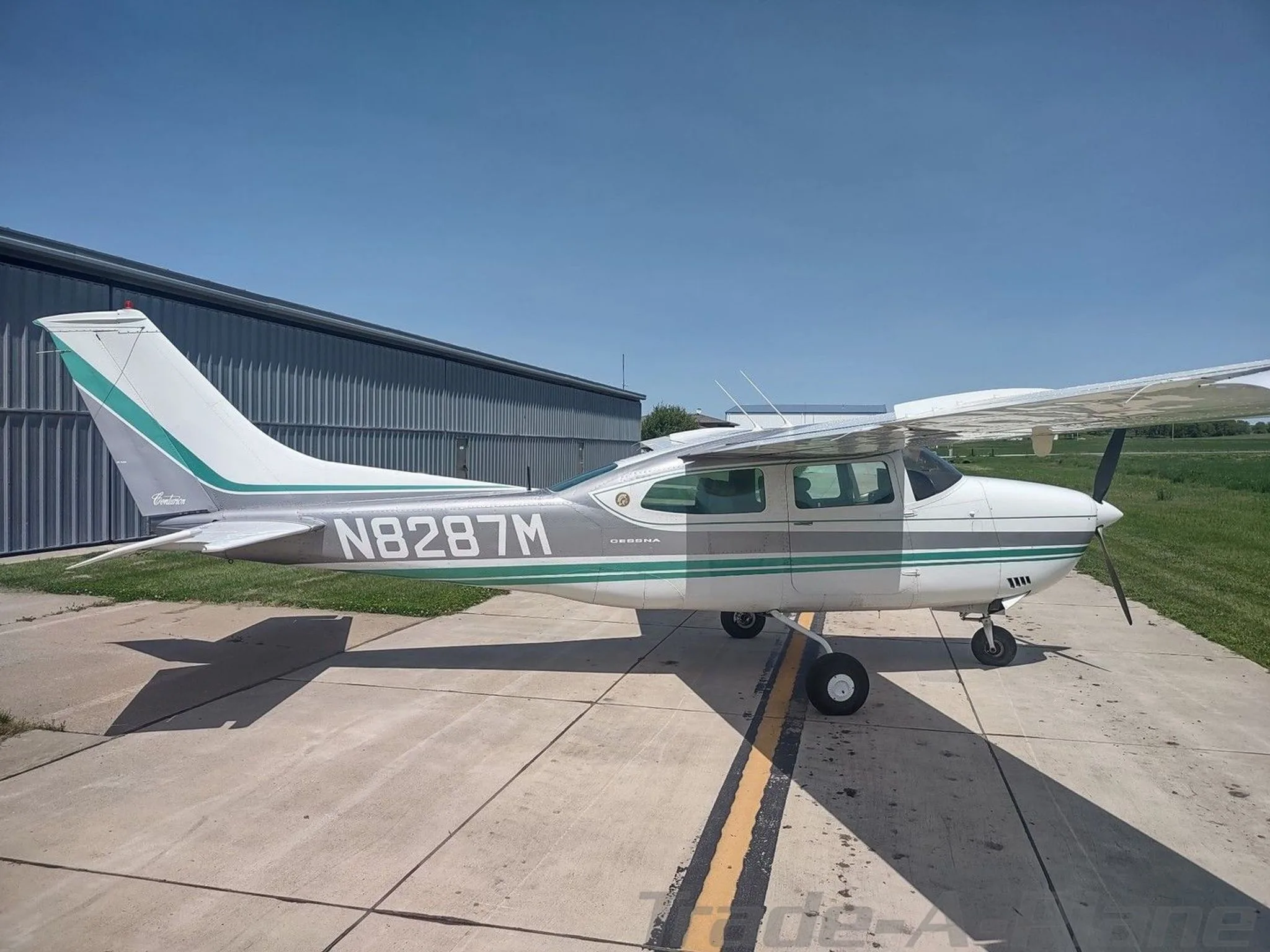 A white and grey Cessna aircraft with green stripes, registration N8287M, parked on a tarmac next to a hangar.