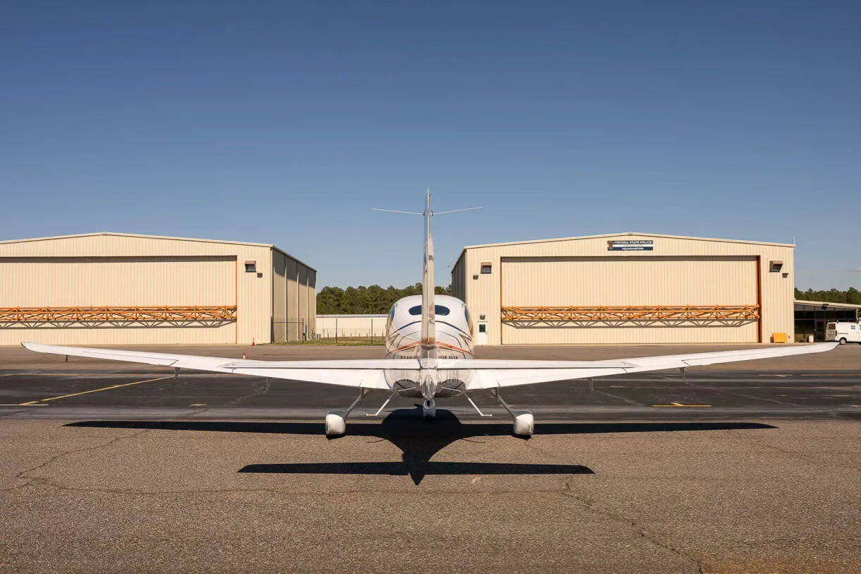 A rear view of a white Cirrus SR22 aircraft with hangars in the background, parked on an airfield.