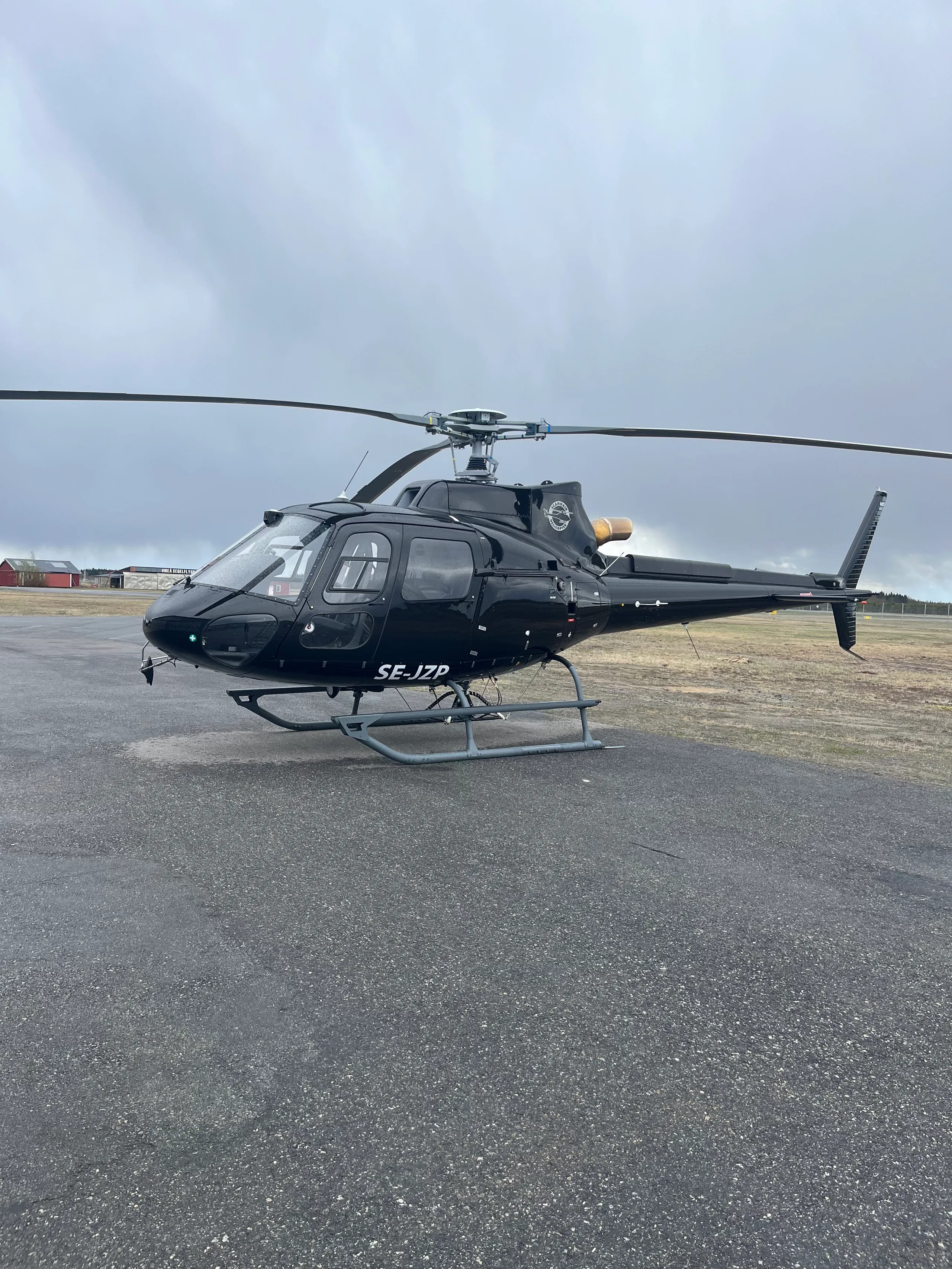 A black Airbus H125 helicopter (SE-JZP) is parked on an asphalt tarmac under a cloudy sky, with buildings in the background.