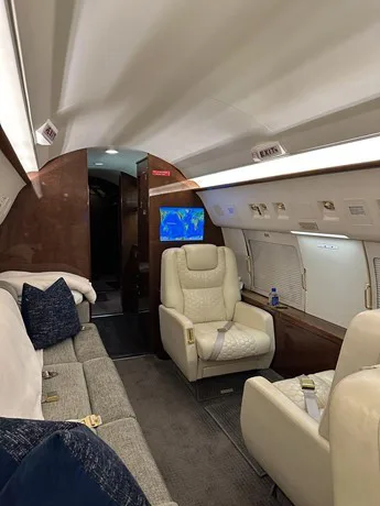 The private jet's cabin features a grey sofa, cream individual seats, and a screen displaying a world map.