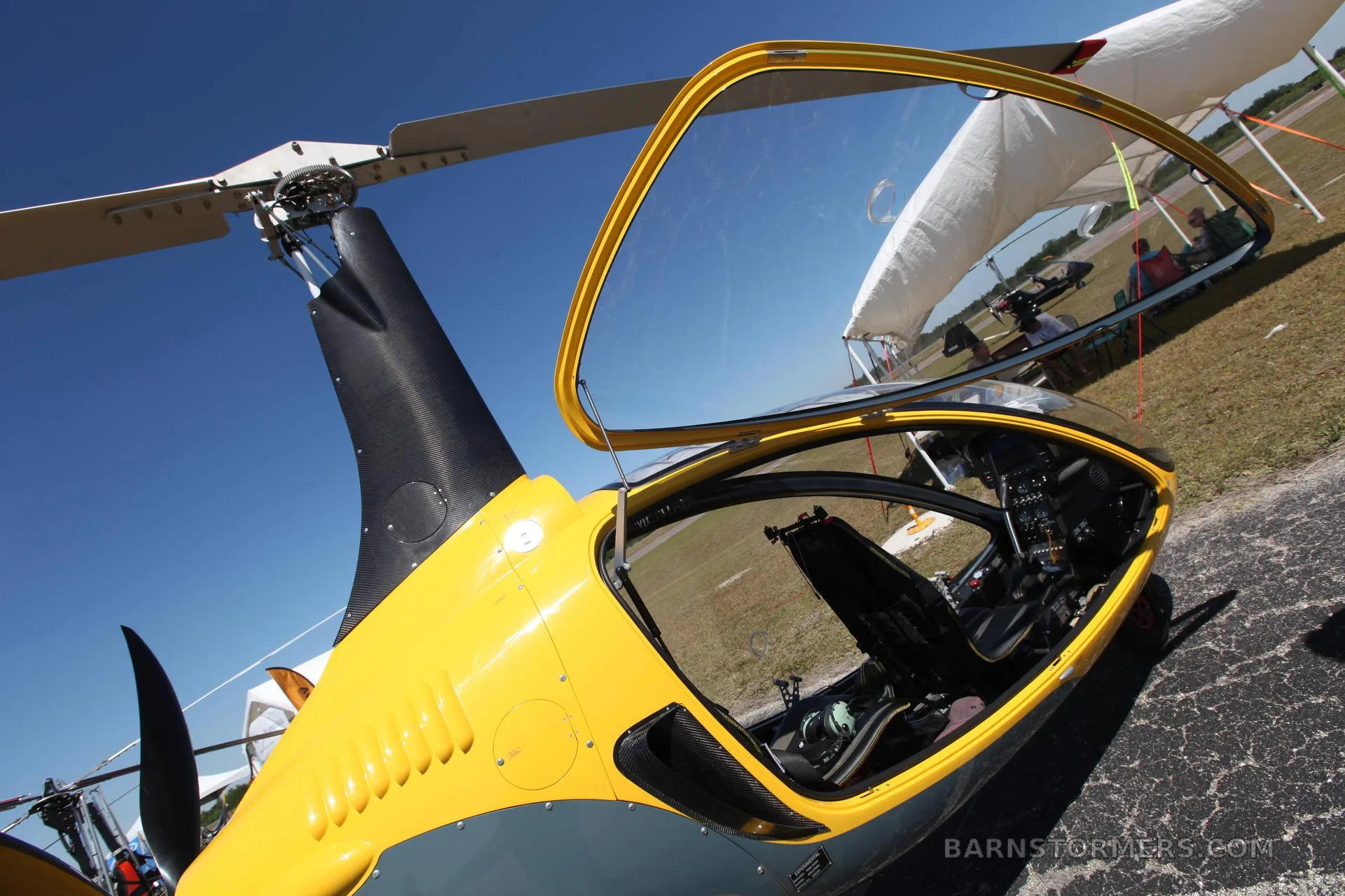 A yellow and grey gyrocopter with its canopy open, parked on an asphalt surface under a clear blue sky.