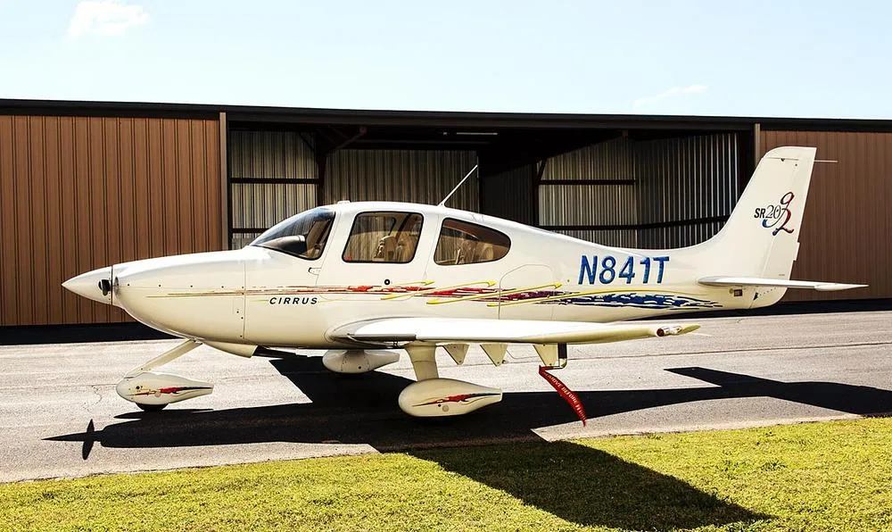 A white Cirrus SR20 aircraft with blue and red accent stripes parked next to a hangar.