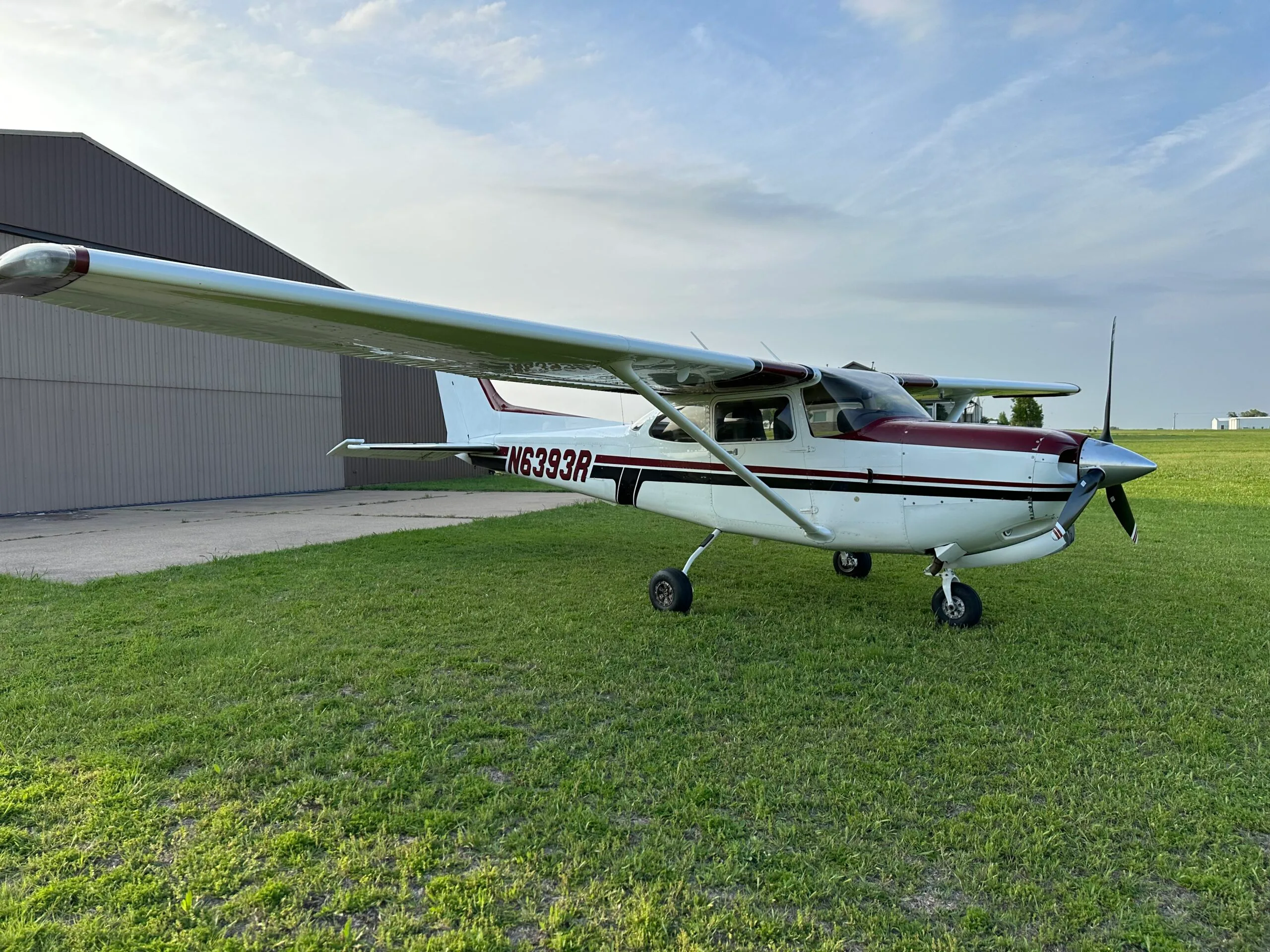 A white and maroon single-engine propeller aircraft, N6393R, is parked on grass next to a hangar under a cloudy sky.
