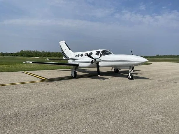 A white twin-engine propeller aircraft with black stripes on the tail and engine nacelles, parked on an asphalt tarmac under a partly cloudy sky.