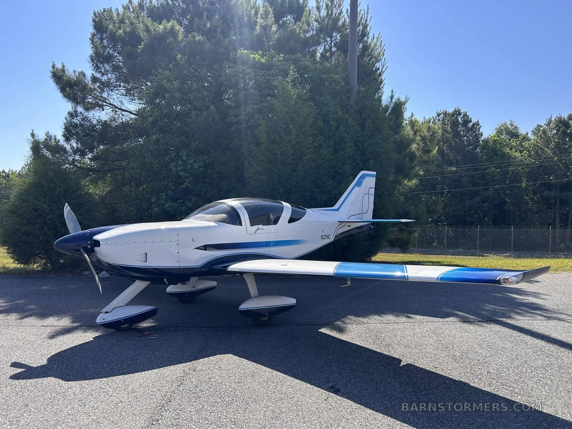 A white and blue single-engine aircraft with registration N37NC is parked on an asphalt surface next to green trees, viewed from the side.