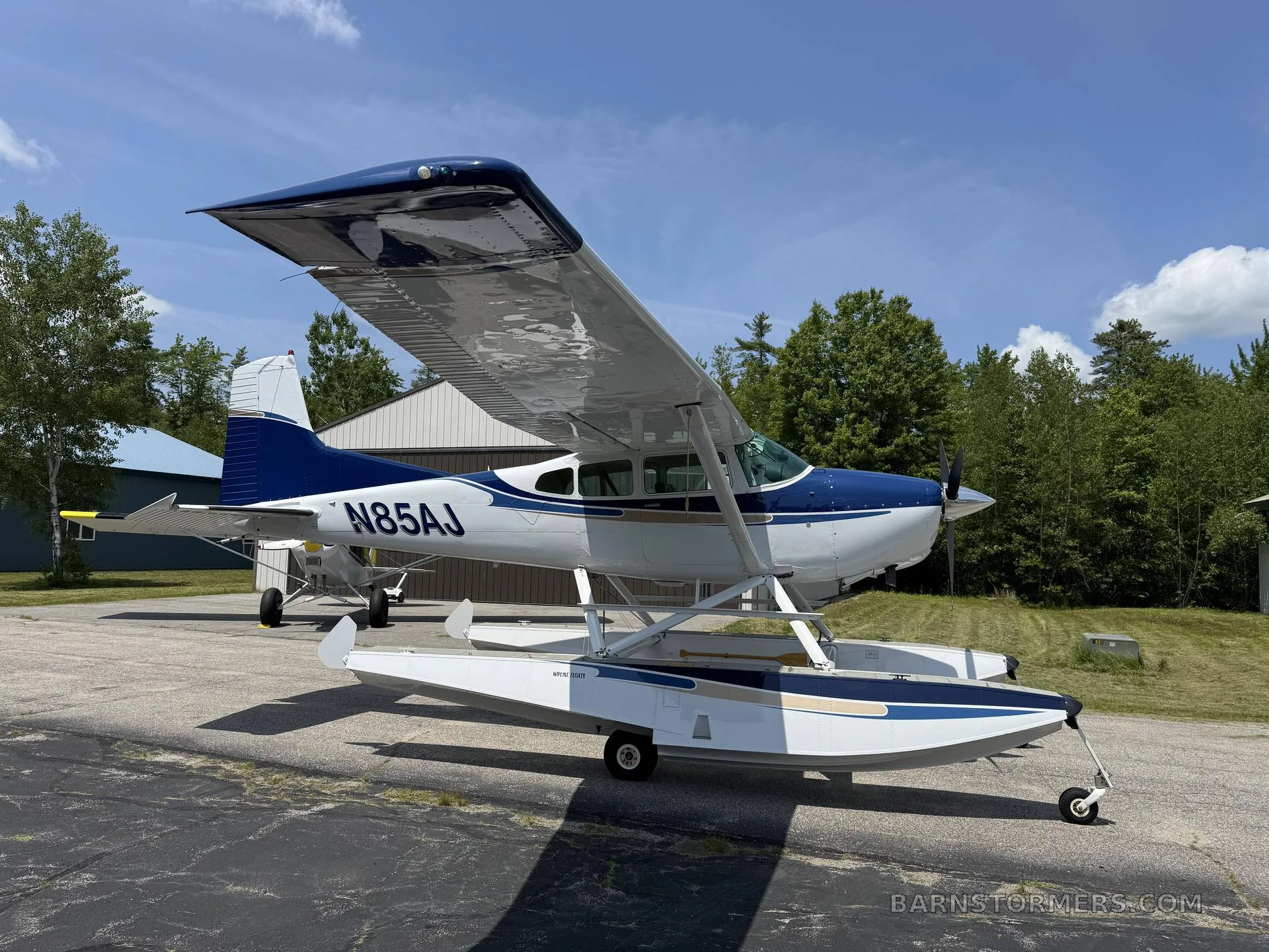 A blue and white floatplane (N85AJ) is parked on an asphalt ramp with trees and buildings in the background.