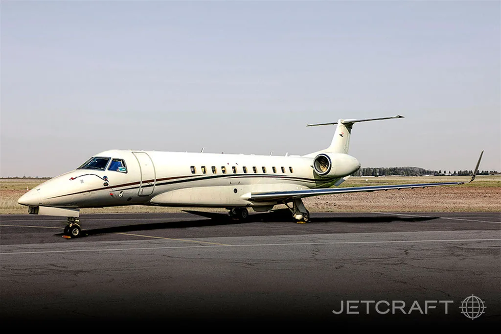 A white Embraer Legacy business jet with a dark stripe parked on an asphalt runway under a clear sky.