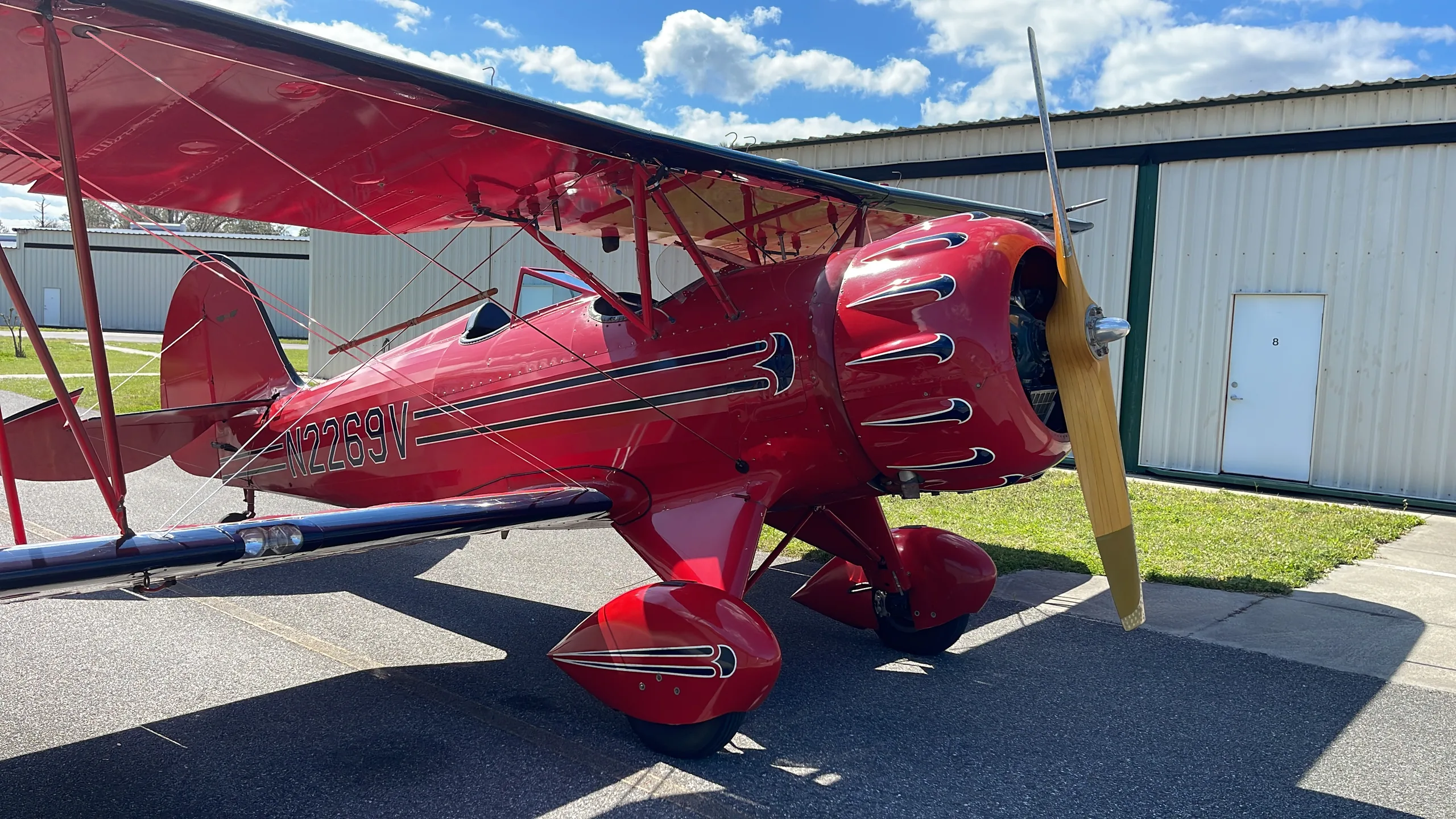 A red biplane with registration N2269V is parked on an asphalt apron next to a white hangar under a partly cloudy sky.