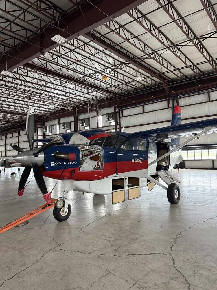 A Kodiak 100 aircraft with blue, red, and white paint scheme, parked in a hangar with engine and cargo bay panels open.