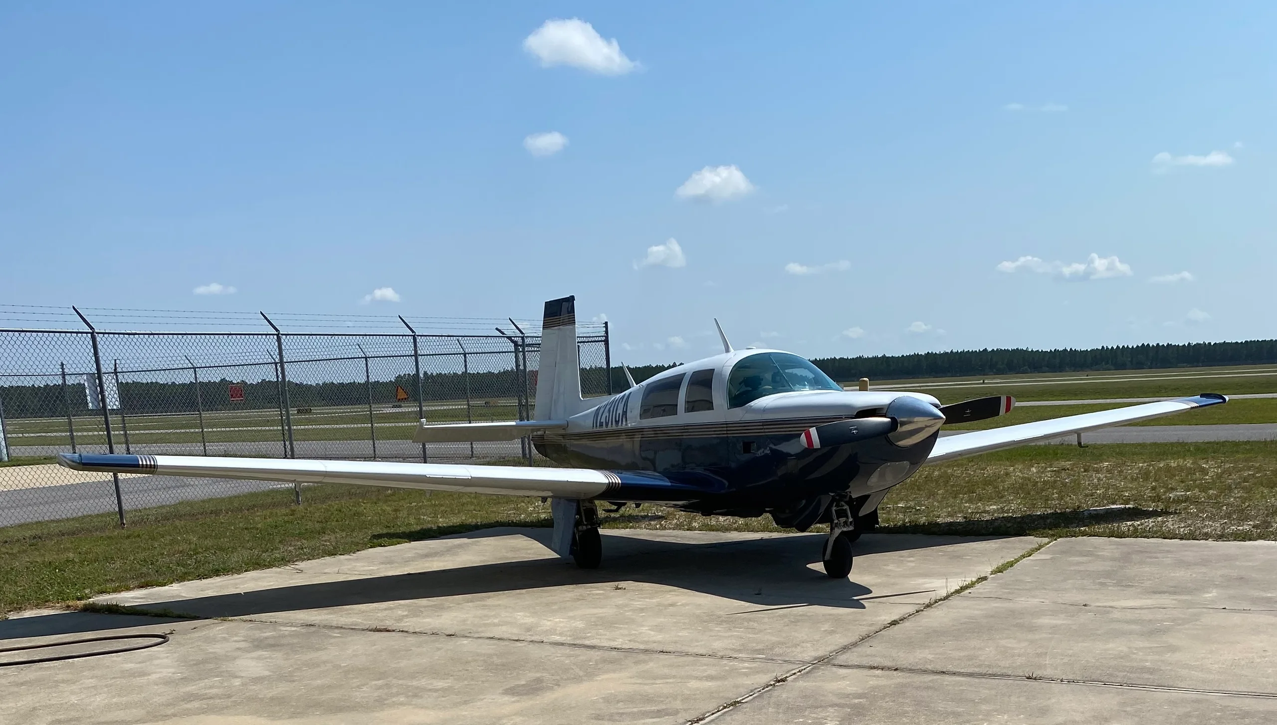 A blue and white single-engine aircraft (N231CA) parked on an airfield with a fence and trees in the background.