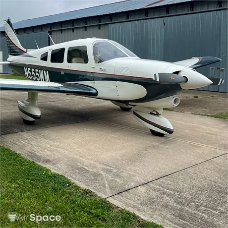 A white and dark green Piper Dakota aircraft, registration N555WM, parked on a concrete apron with a hangar in the background.