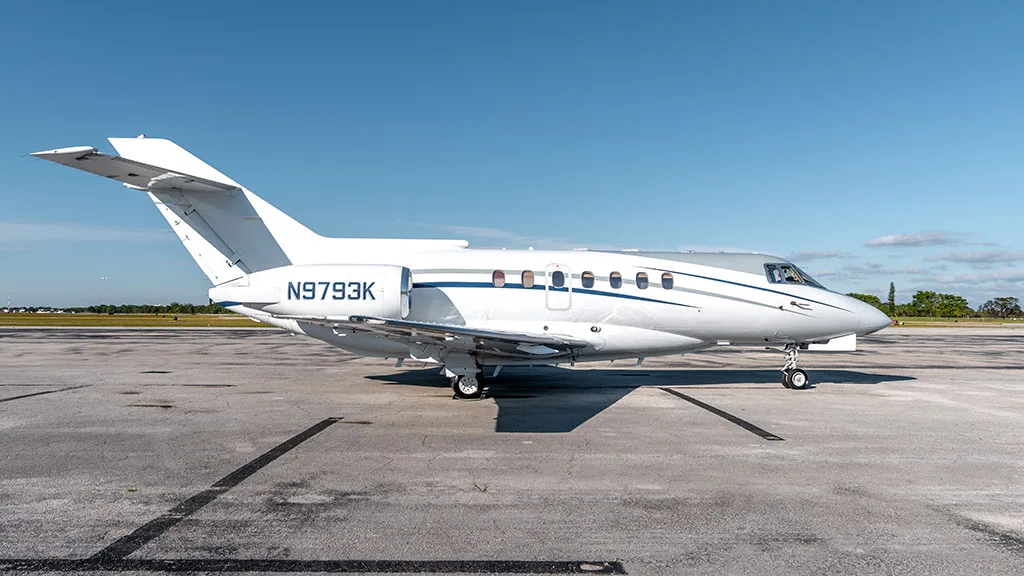 A white business jet with blue and grey stripes, tail number N9793K, parked on an airfield from a side view.