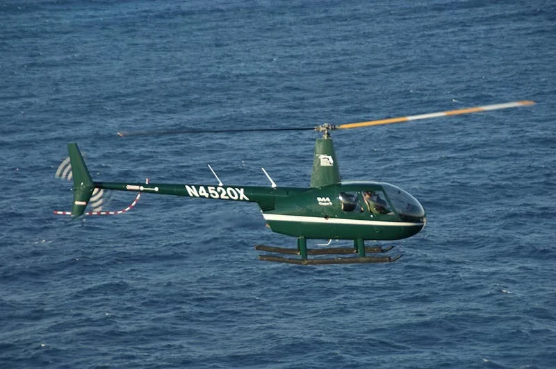 A dark green Robinson R44 Clipper II helicopter, registration N4520X, flies low over blue ocean water.