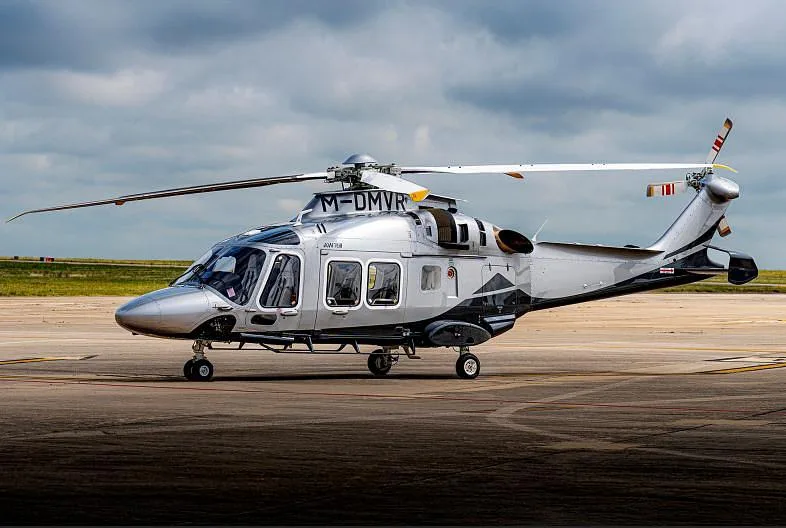 A silver AgustaWestland AW139 helicopter with black and grey accents sits on an airport tarmac under a cloudy sky.