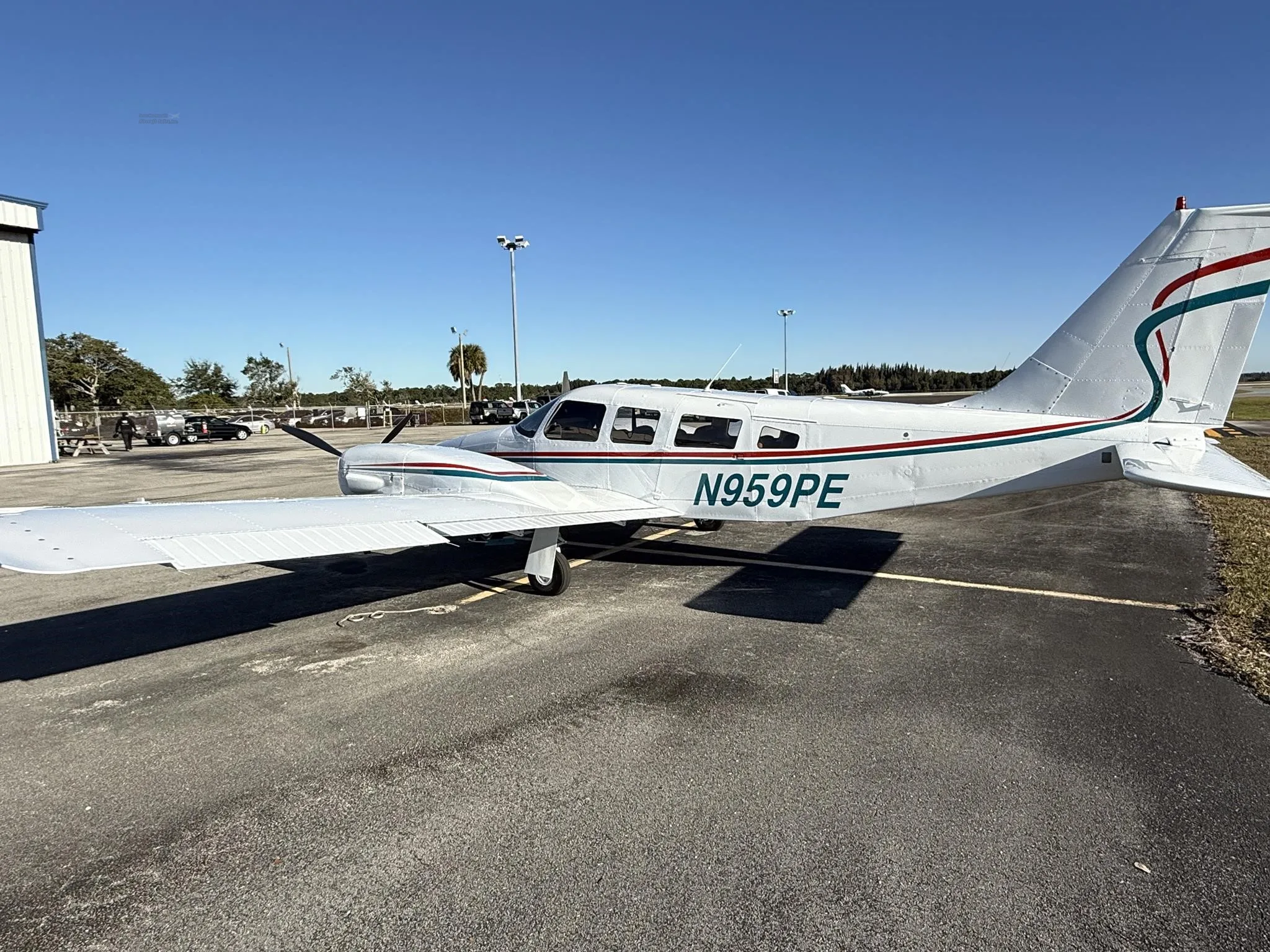 A white twin-engine Piper Seneca II, N959PE, with teal and red stripes, parked on an airport tarmac next to a hangar.