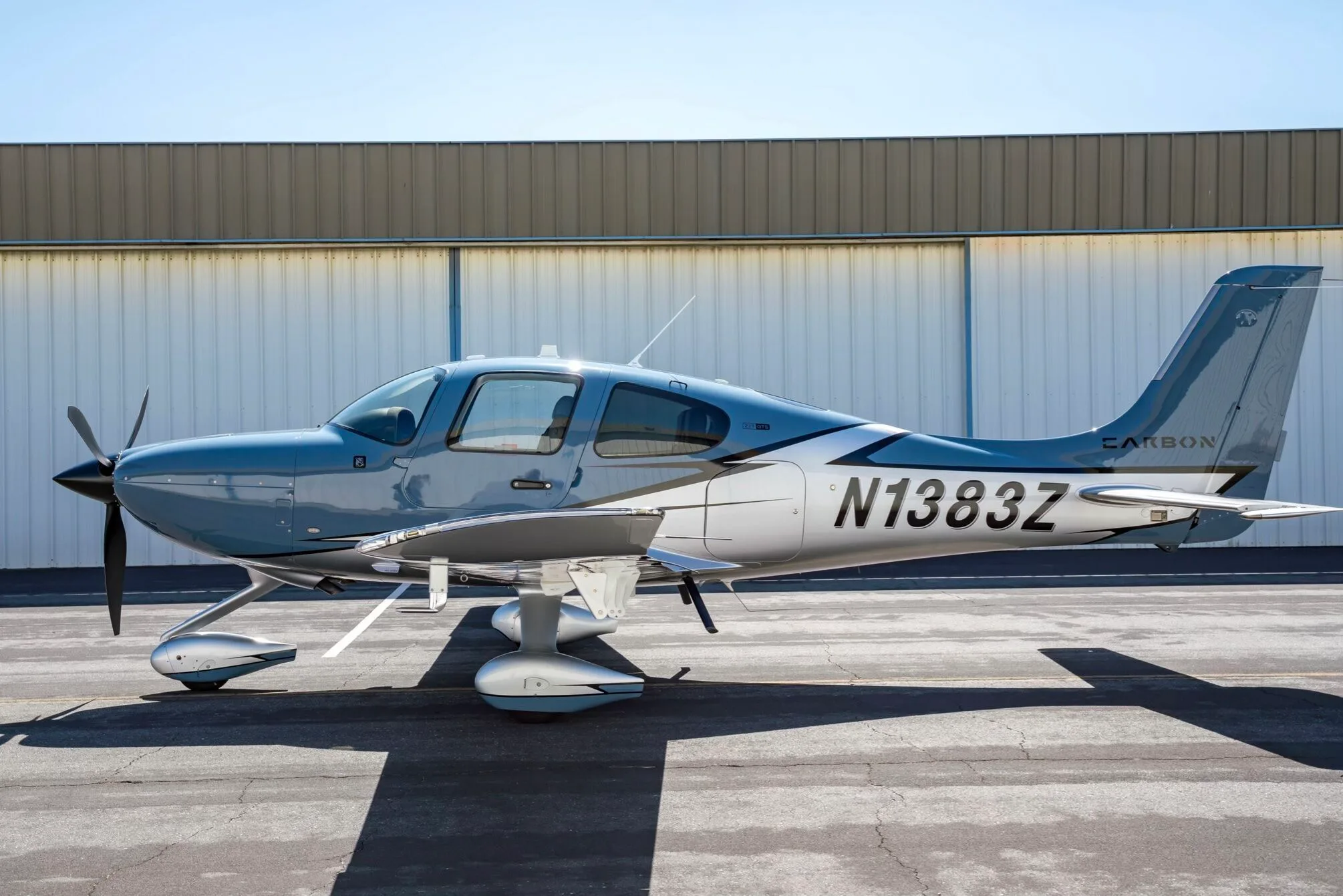 A light blue and silver Cirrus SR22T aircraft, N1383Z, parked on an tarmac in front of a hangar.