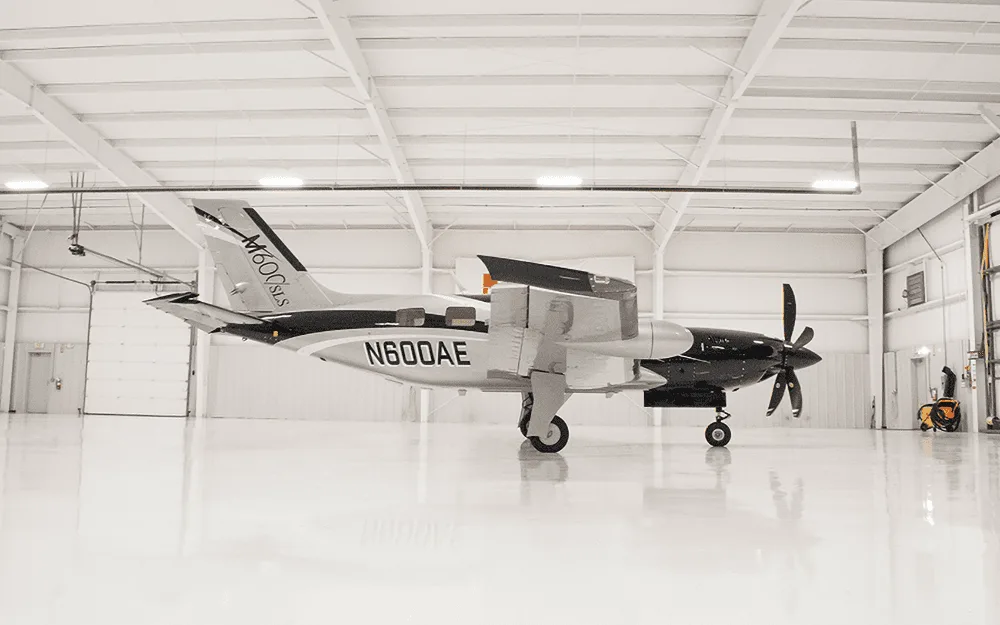 A Piper M600/SLS aircraft with registration N600AE, featuring a black and silver paint scheme, parked inside a brightly lit hangar.
