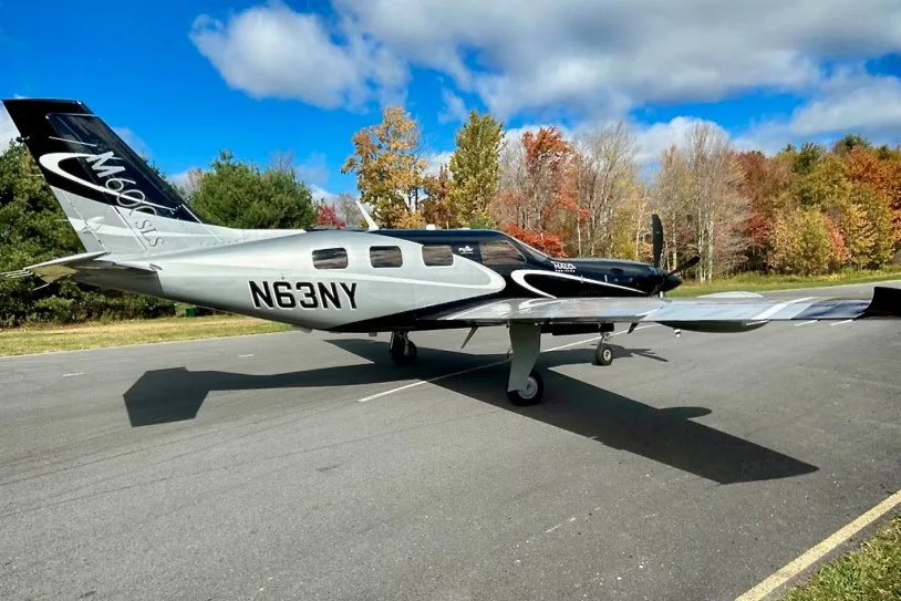 A three-quarter view of a black and silver Piper M600 SLS aircraft (N63NY) on an airfield, with colorful autumn trees behind it.