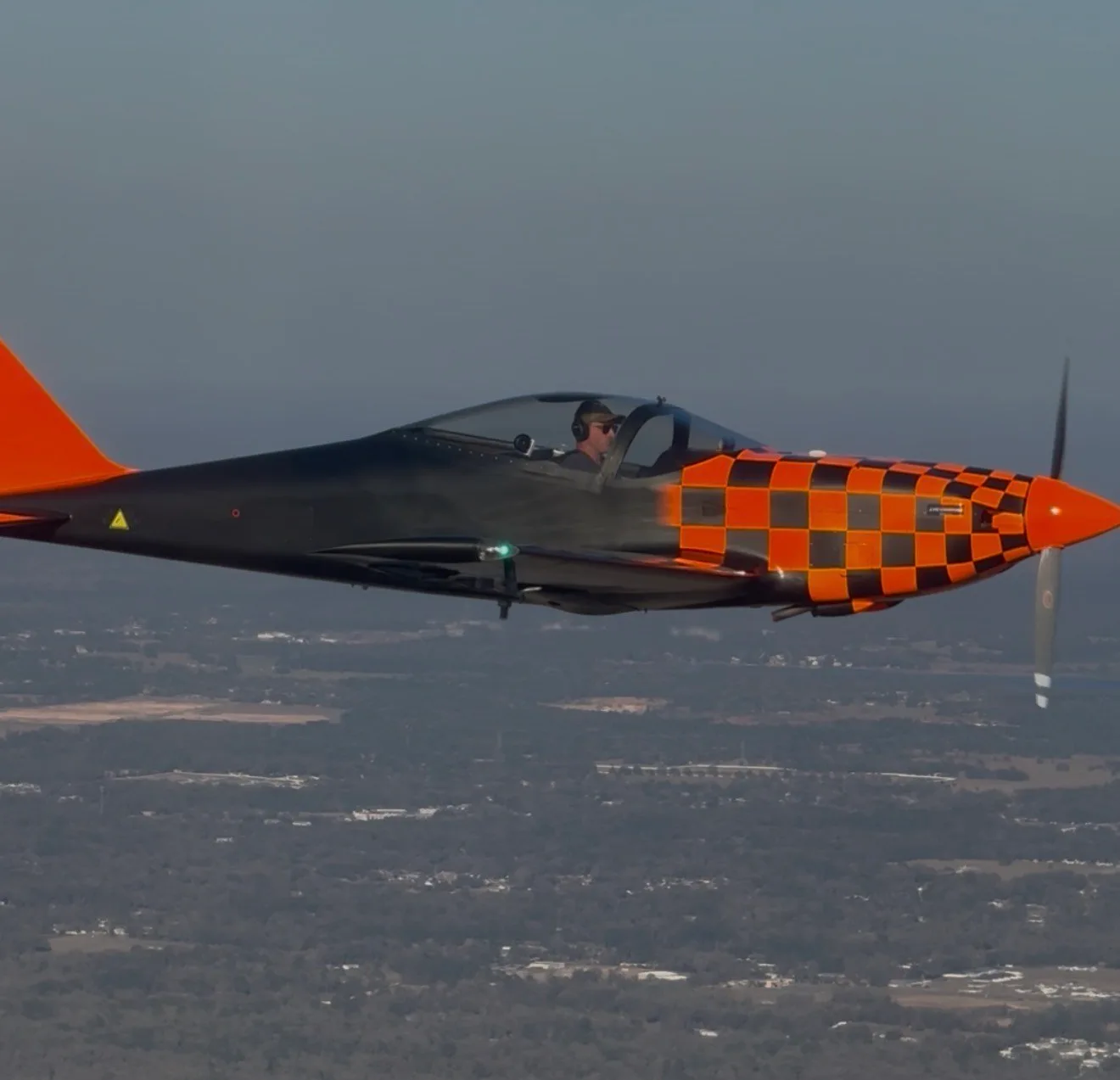 A black and orange checkered experimental aircraft, N7224A, flies against a hazy sky over a landscape.