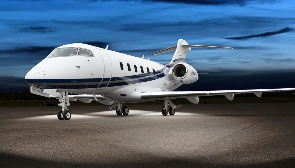 A white Bombardier Challenger business jet with blue stripes, registration N987HP, parked on a tarmac at dusk with its landing lights on.