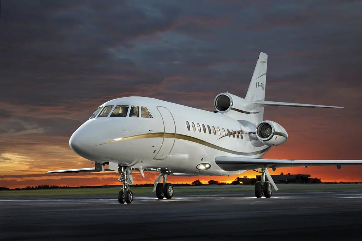 A white Dassault Falcon 900EX with gold stripes, registration XA-FXL, parked on a tarmac at sunset.
