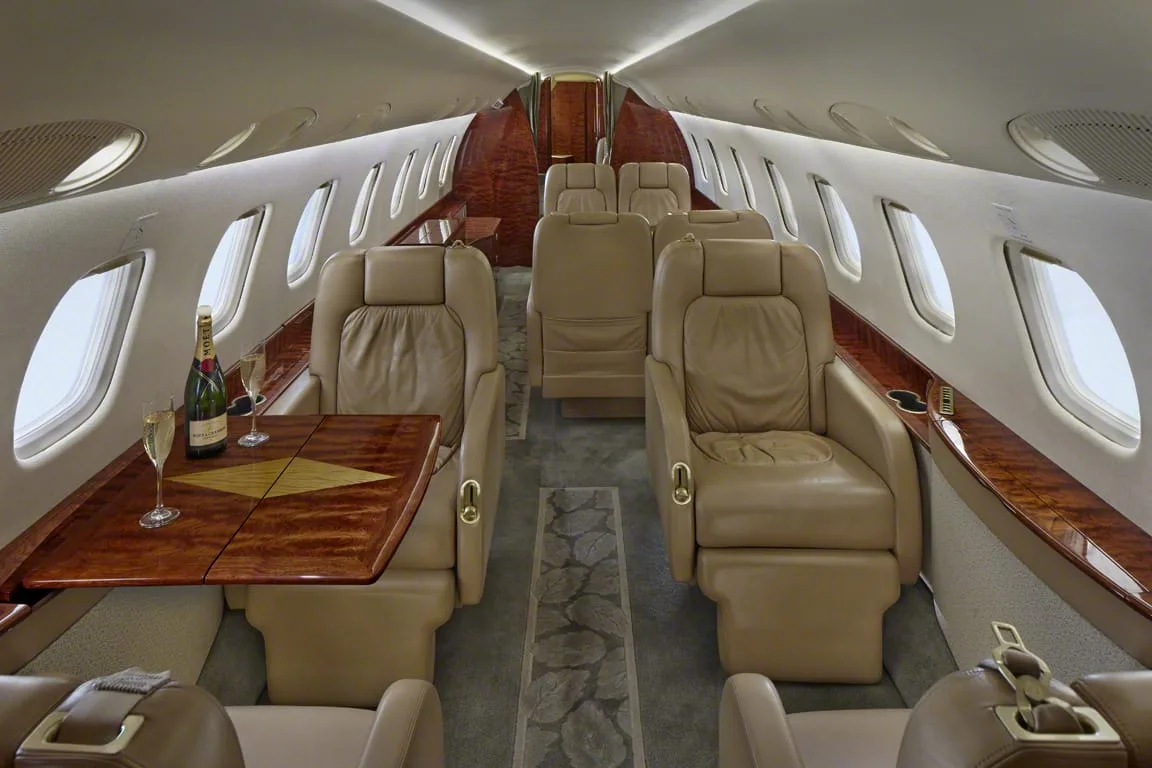 A view down the aisle of a private jet cabin with beige leather seats, a wooden table, and champagne.