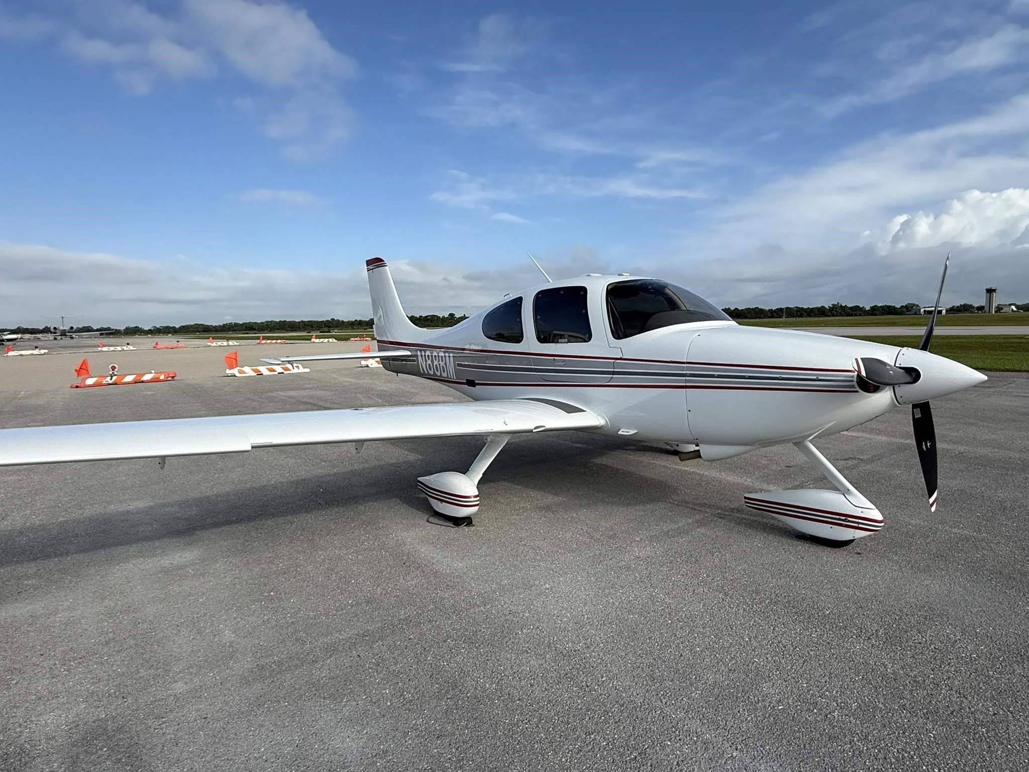 A white Cirrus SR22 aircraft with red and gray stripes, tail number N88BM, parked on an airport tarmac under a partly cloudy sky.