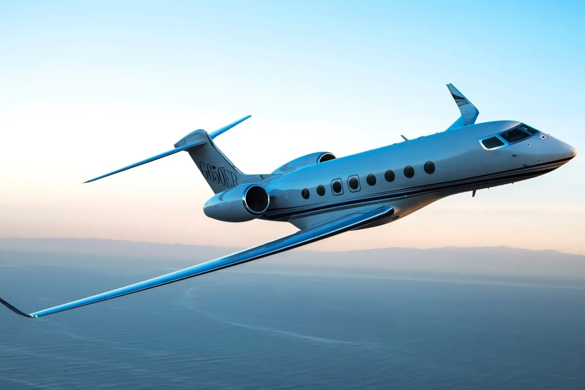 A white and blue Gulfstream G650ER private jet flies over water with a sunset sky in the background.