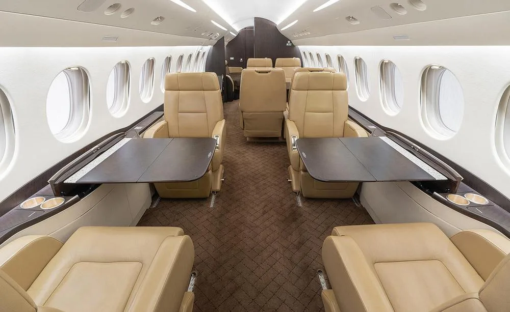 The interior cabin of a business jet features tan leather seats arranged in a club configuration with dark wood tables.
