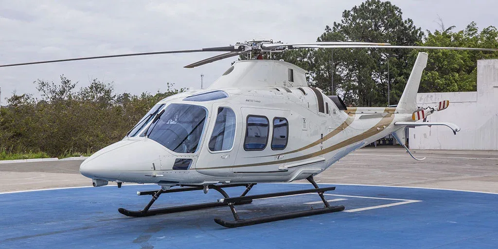A white helicopter with gold stripes is parked on a blue helipad, viewed from the side with trees in the background.
