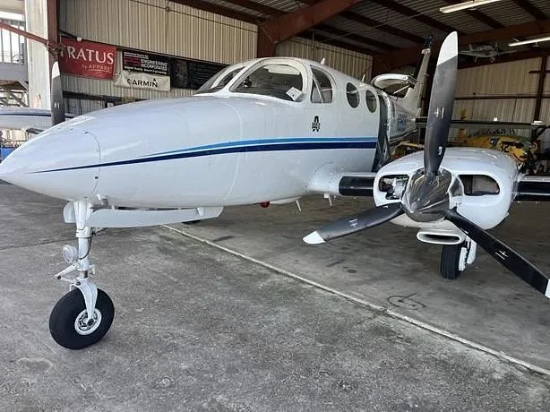 A white twin-engine propeller aircraft with blue stripes is parked inside a hangar, viewed from the front three-quarter angle.