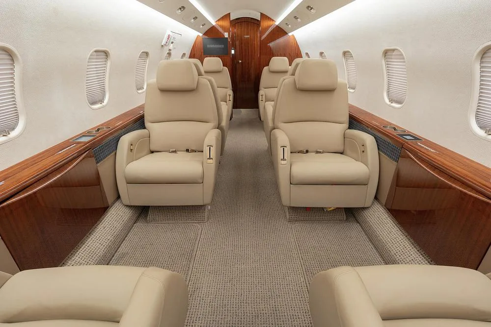 A view down the aisle of a business jet's cabin, showing rows of beige leather seats and dark wood paneling along the walls.