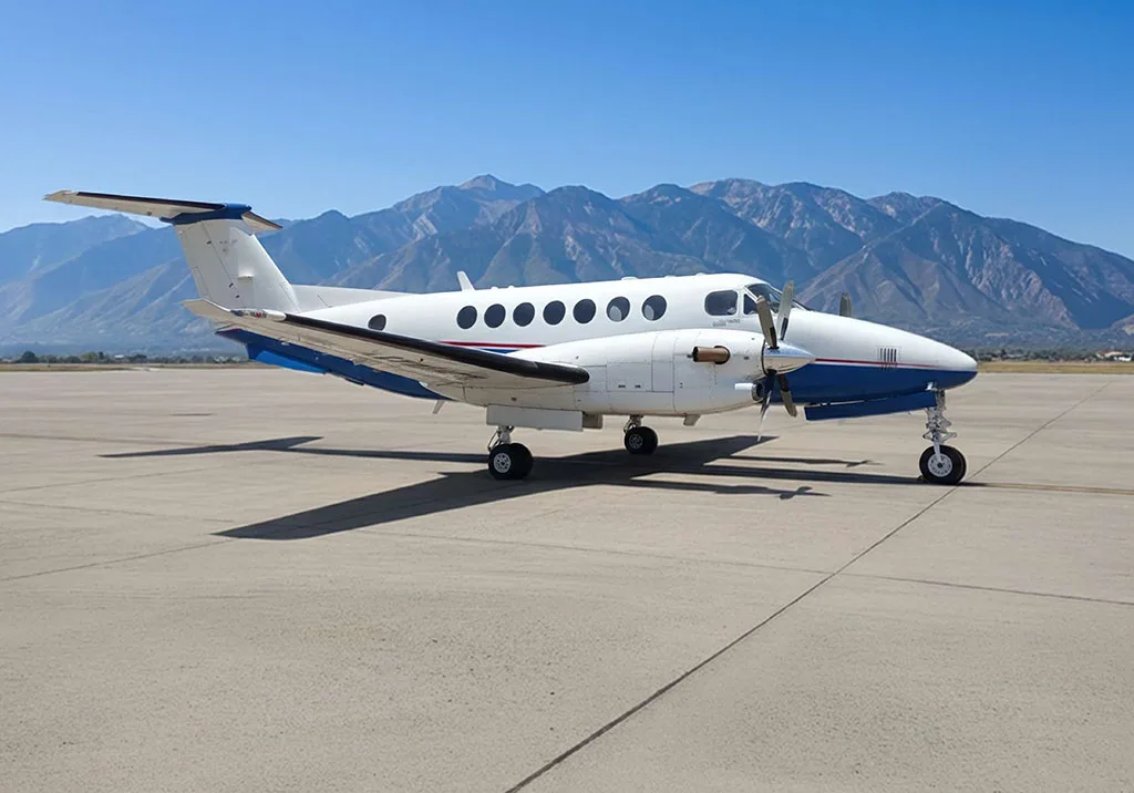 A white and blue turboprop aircraft, likely a King Air, parked on an asphalt runway with mountains in the background under a clear blue sky.