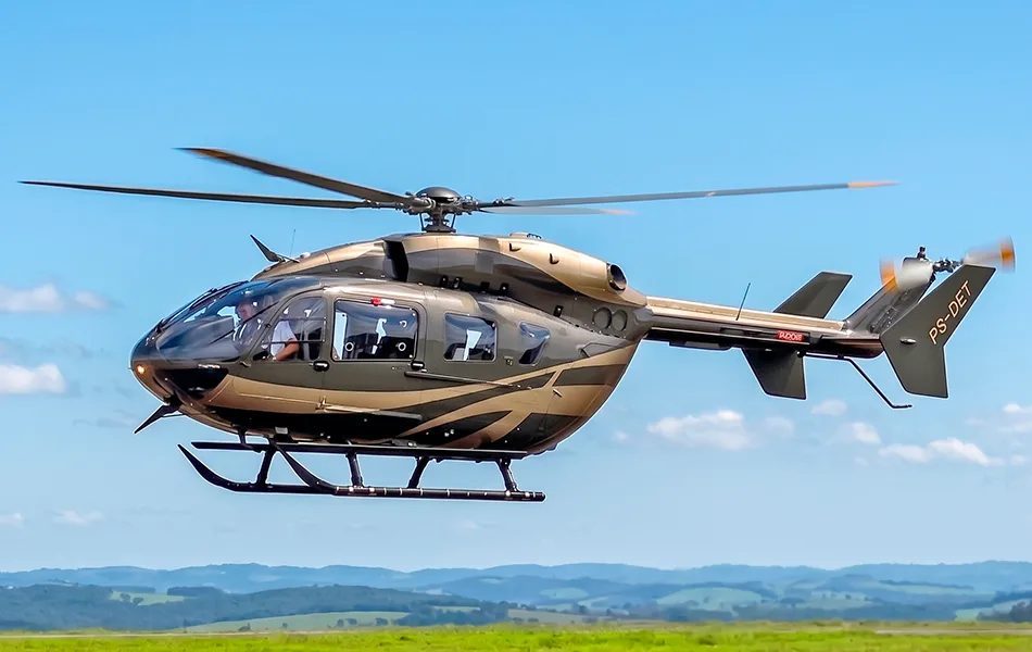 A brown and tan patterned helicopter, registration PS-DET, hovers over a green, hilly landscape under a blue sky.