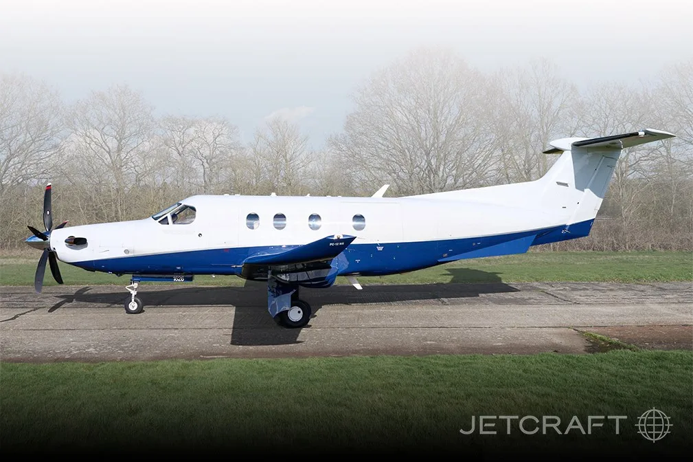 A white and blue Pilatus PC-12 turboprop aircraft parked on a tarmac with trees in the background.