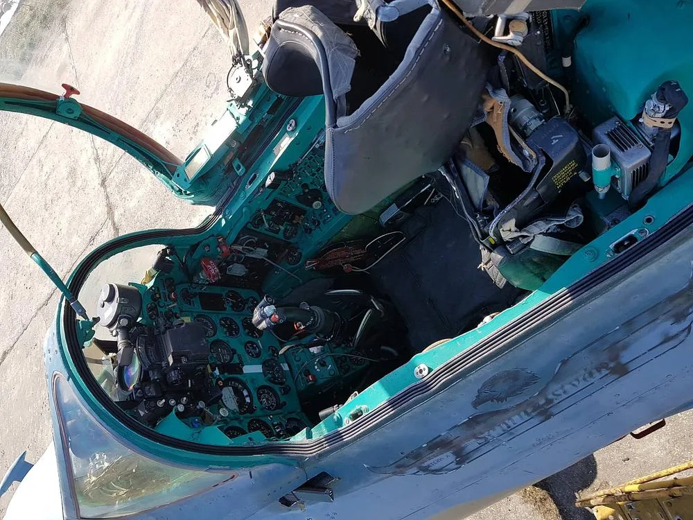 An aerial view looking down into the open cockpit of a fighter jet, revealing the green instrument panel and ejection seat.