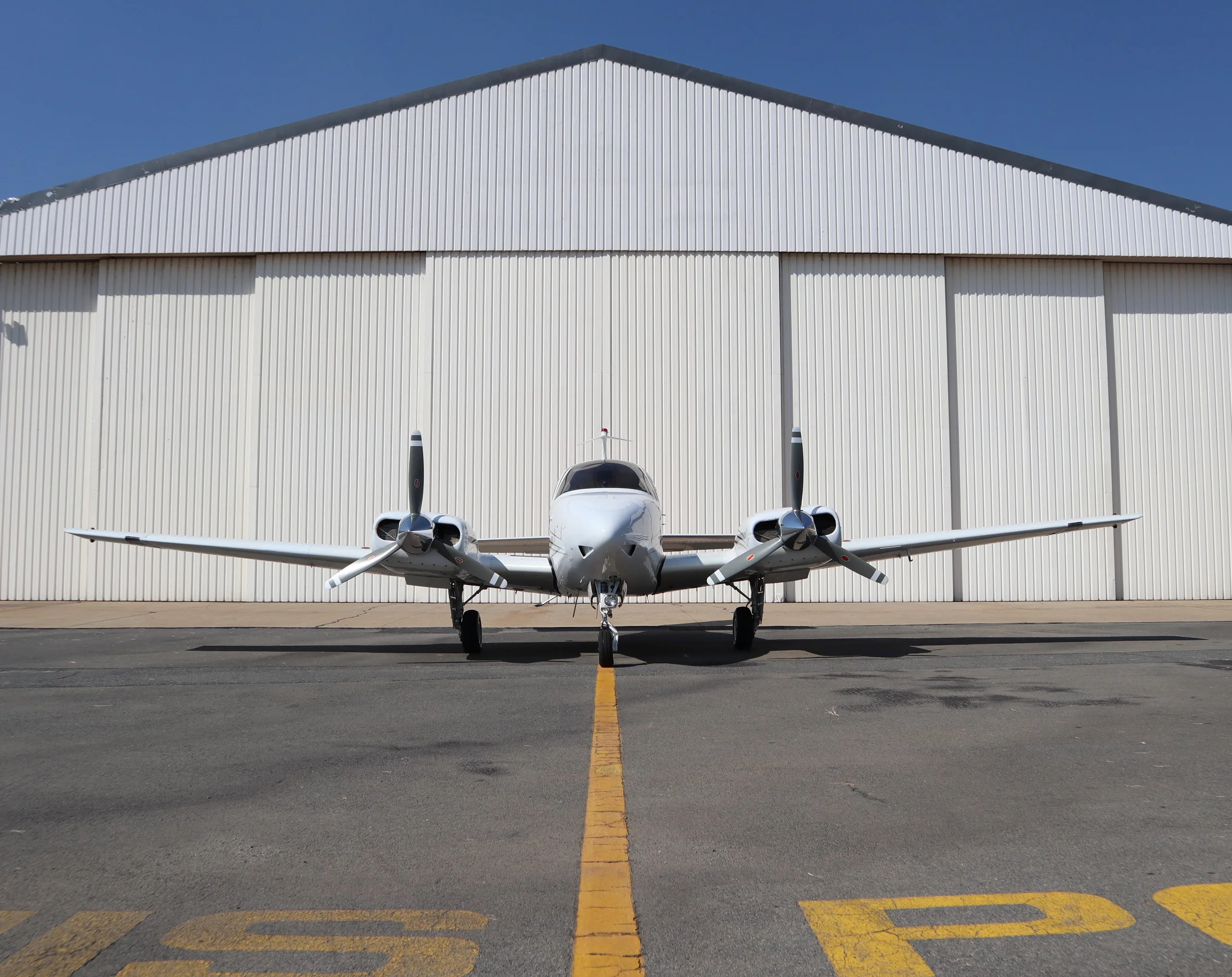 A white twin-engine propeller aircraft is parked on an asphalt apron in front of a large white hangar under a clear blue sky.