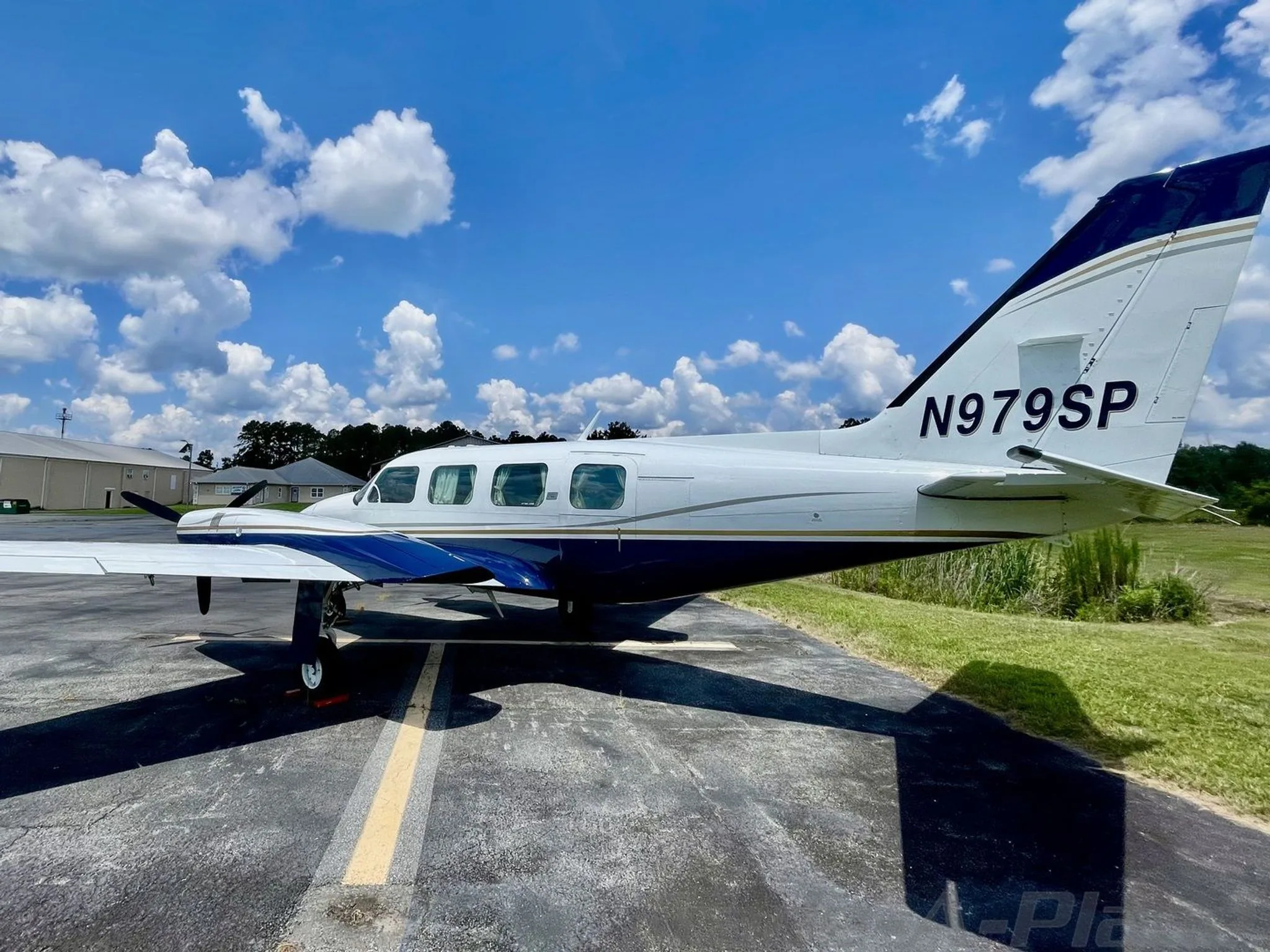 The rear-side view of the white and blue Piper aircraft, N979SP, parked on an asphalt ramp with grass and buildings in the background.