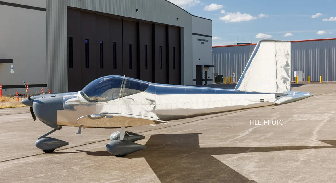 A polished silver and grey single-engine aircraft with a bubble canopy parked on an airfield in front of a hangar.