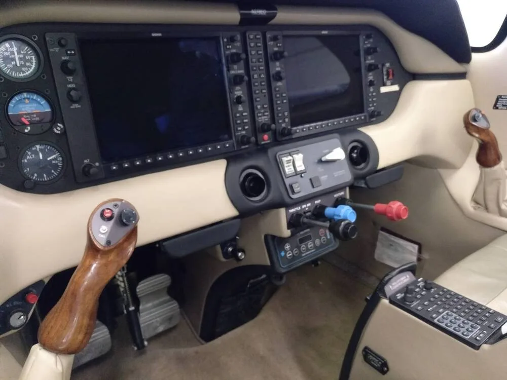 The cockpit of an aircraft featuring two large display screens, traditional gauges, and a wooden side-stick control.