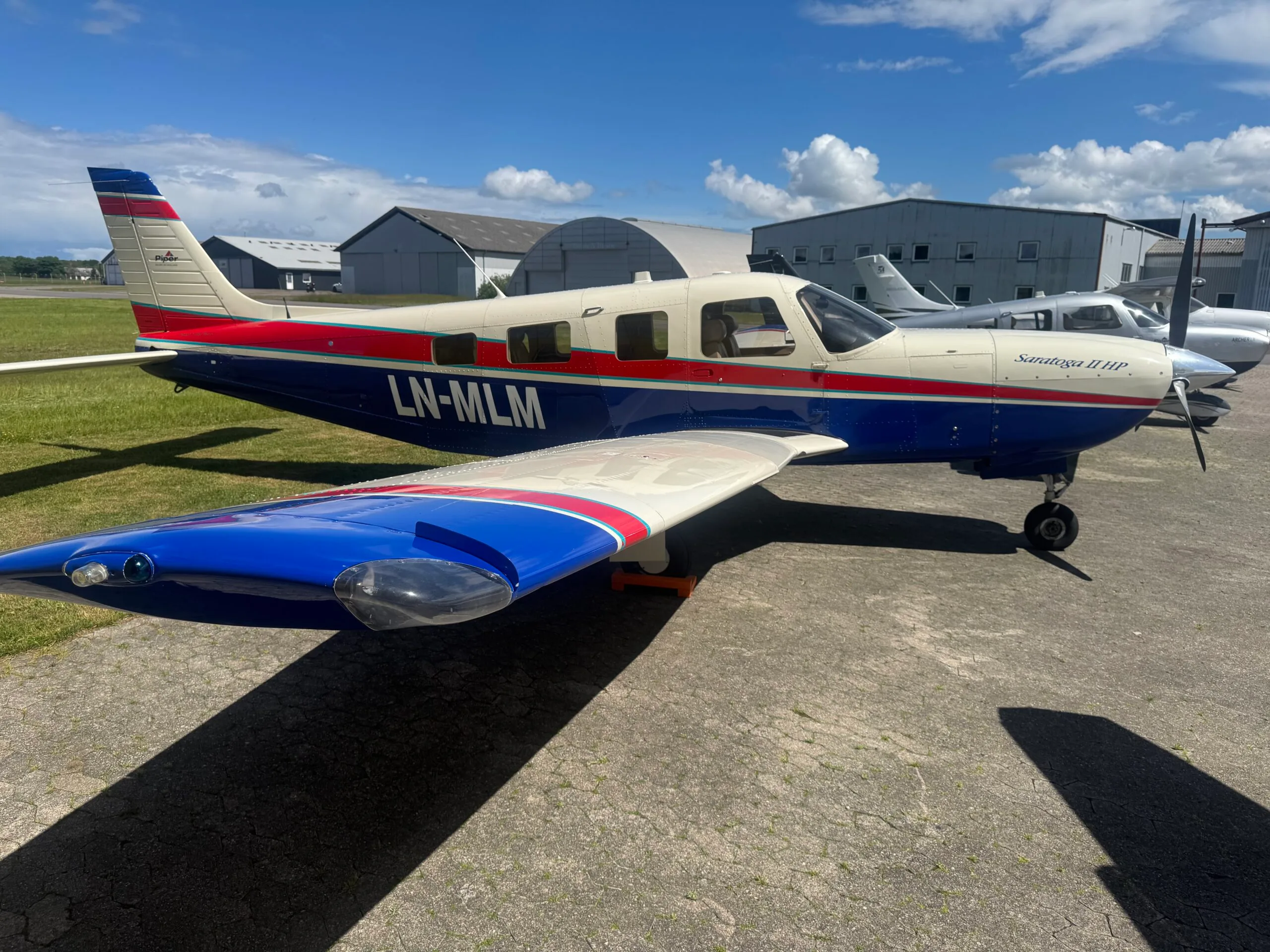 A white, blue, and red Piper Saratoga II HP aircraft with registration LN-MLM parked on an airfield next to hangars.