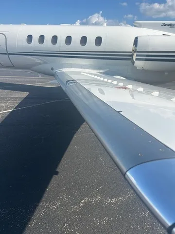 A close-up view of the wing and mid-fuselage of a white business jet with dark stripes, showing the engine nacelle in the background.