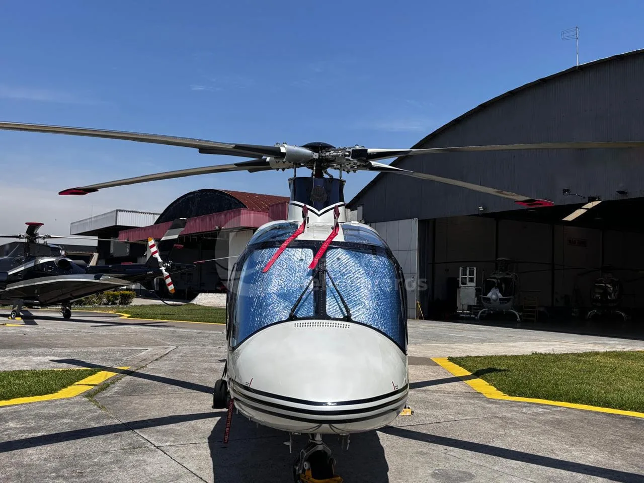 A head-on view of a white and black Agusta A109 Power helicopter with a sunshade, parked on an airfield.