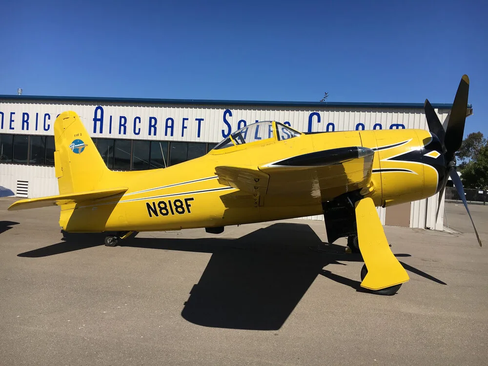 A yellow Grumman F8F Bearcat aircraft with black accents, registration N818F, parked on an asphalt tarmac in front of a hangar.