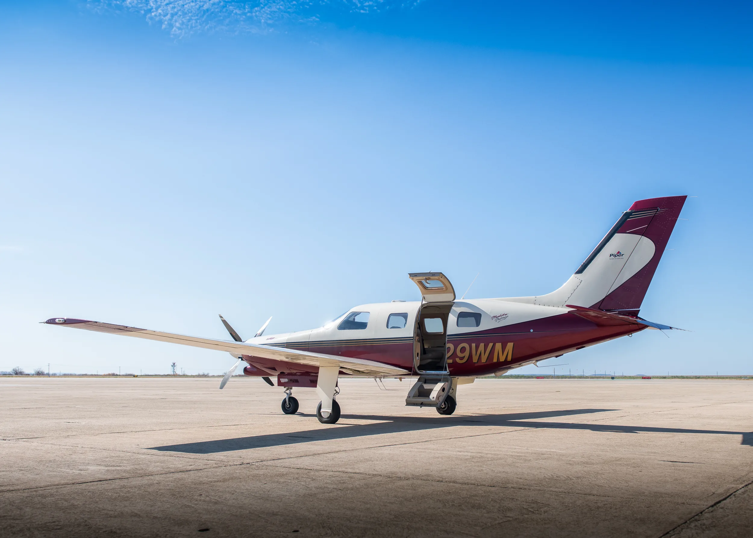 A three-quarter front view of the maroon and white Piper M350 aircraft (N529WM) with its cabin door open, parked on an airfield.