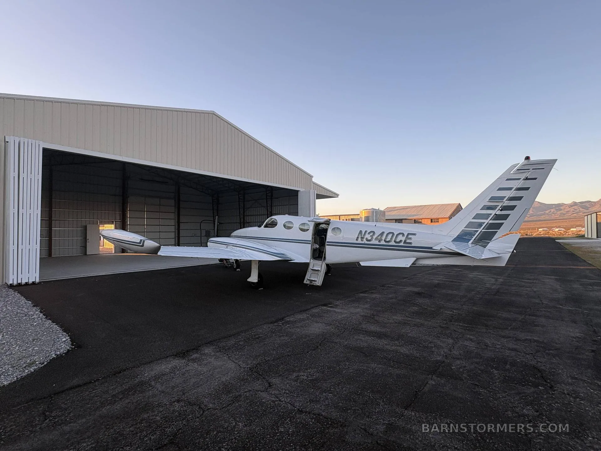 A white Cessna 340 (N340CE) with blue stripes parked outside a hangar at sunset.