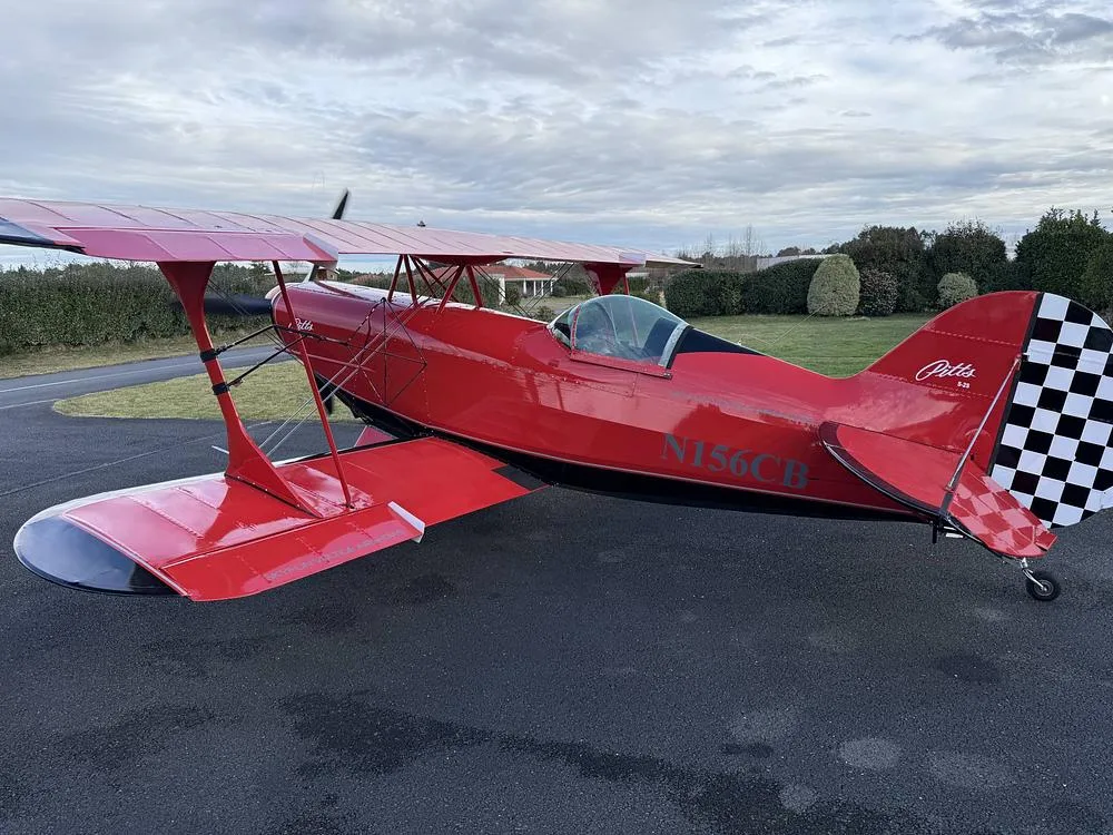 A red Pitts S-2S biplane with black accents and a checkered tail, registration N156CB, parked on an asphalt surface under a cloudy sky.