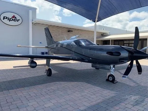 A dark grey Piper M600/SLS turboprop aircraft parked on a paved area in front of a building with a "Piper" logo.