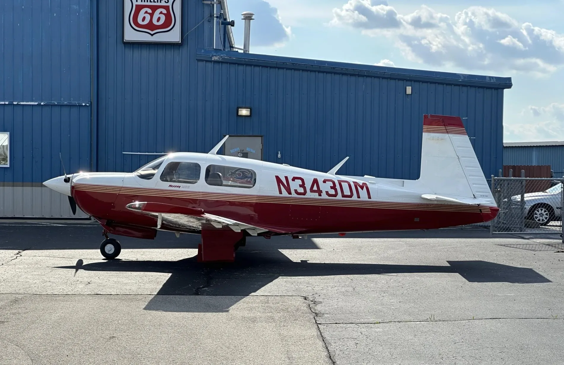 A full side view of a red and white Mooney aircraft (N343DM) parked in front of a blue hangar with a Phillips 66 sign.