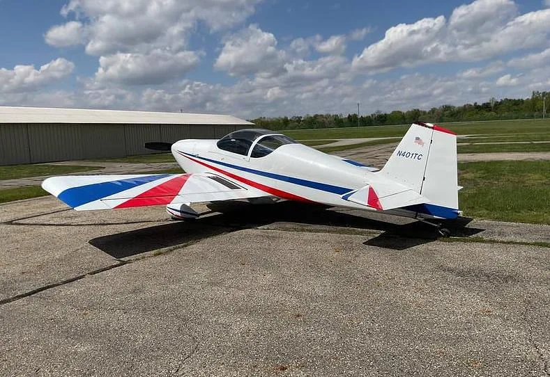 A white single-engine aircraft with red and blue stripes on the fuselage and wings, parked on a tarmac with a hangar in the background.